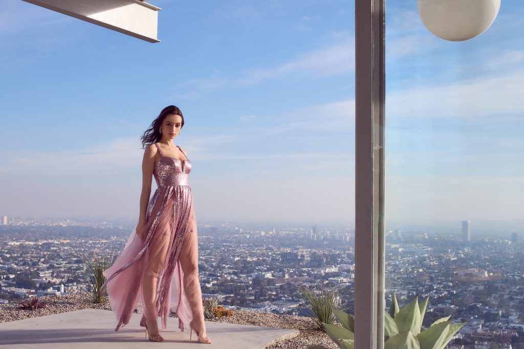 A woman in Roxanne Light Pink Gladiator Gown standing on a balcony overlooking a city.